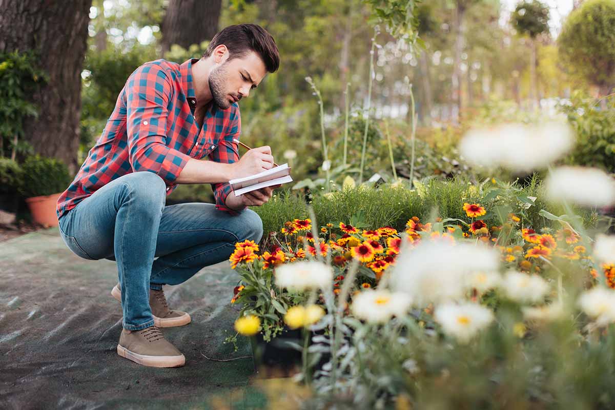 A young man bending down over a flower bed, holding a pen and writing in a notebook, with a garden scene in soft focus in the background.