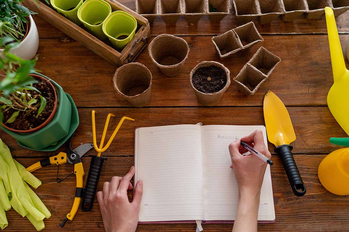 A close up of an open notebook with a hand holding a pen making notes. Surrounding the book are garden supplies, set on a wooden surface.