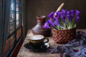 A close up horizontal image of purple crocus flowers growing in a pot set on a rustic wooden table near a window.