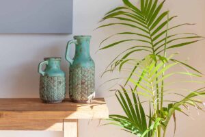 A close up horizontal image of a tall houseplant growing next to a wooden sideboard with two light blue decorative vases.