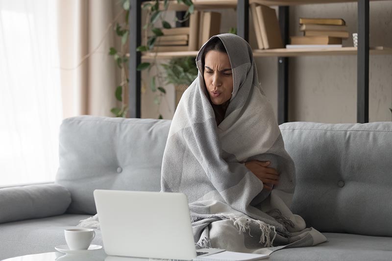 A horizontal image of a woman sitting on the couch wrapped in a blanket watching a laptop computer during the winter.