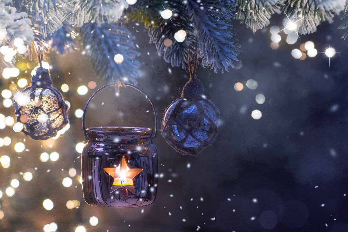 A close up horizontal image of a Christmas lantern and other decorations hanging from a tree with sparkly lights in soft focus in the background.