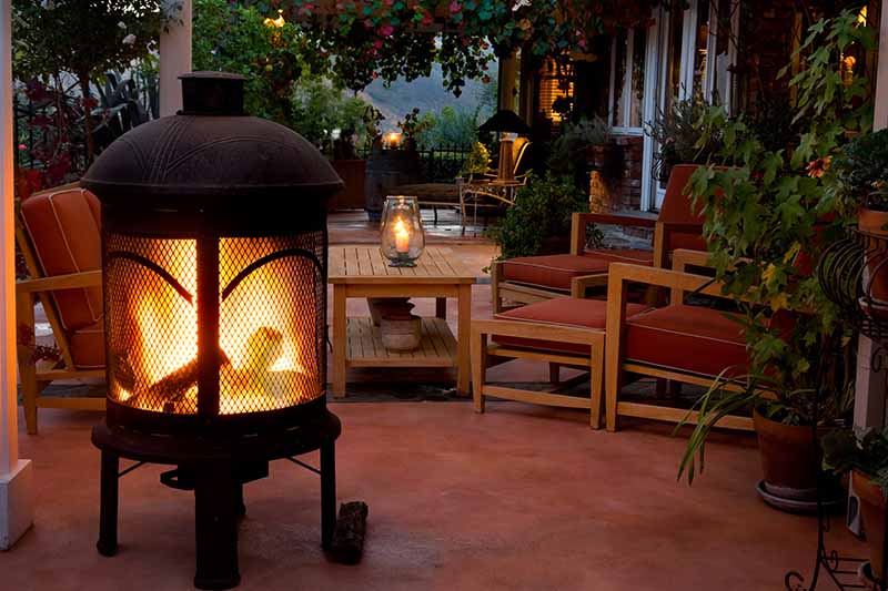 A horizontal image of an outdoor living space with wooden furniture, candles and a large patio heater burning wooden logs.
