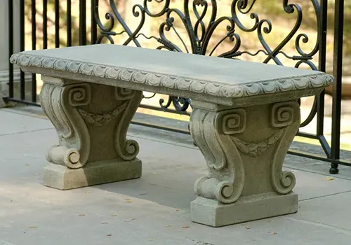 A close up of a rectangular cast stone garden bench stet on a tiled patio with wrought iron in the background.
