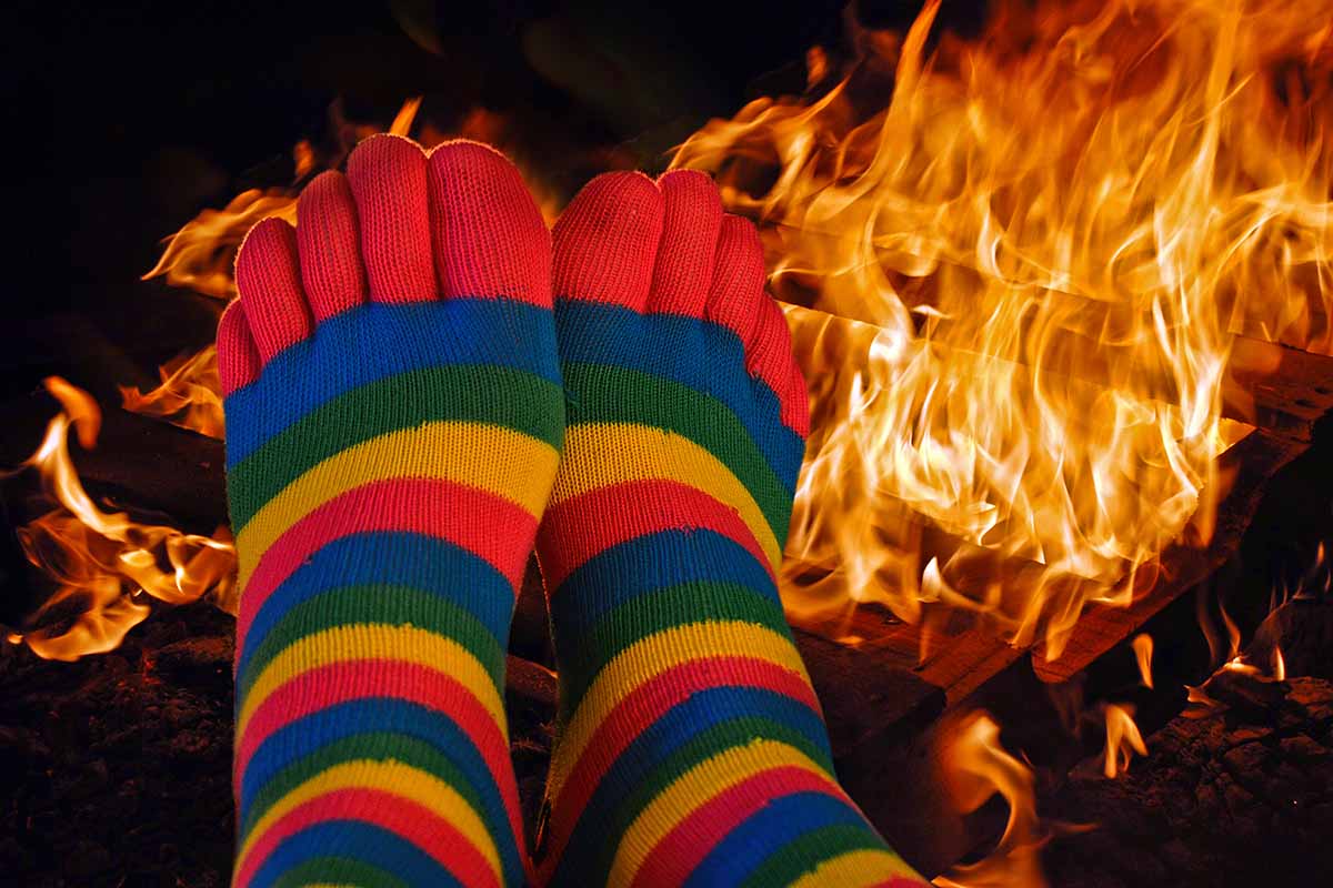 A close up horizontal image of feet with colorful socks warming in front of a large outdoor fire.