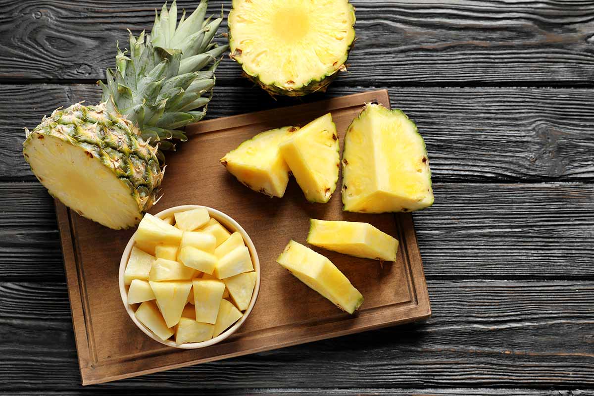 A close up horizontal image of pieces and slices of pineapple set on a wooden chopping board on a wooden surface.