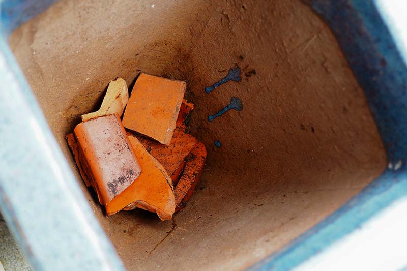 A close up top down image of an empty container with broken pottery pieces placed in the bottom to help with drainage.