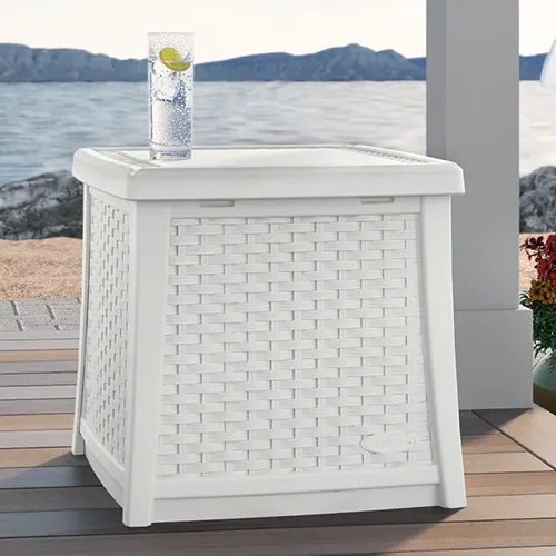A close up square image of a white storage box that doubles as a side table on a wooden deck with the ocean in the background.