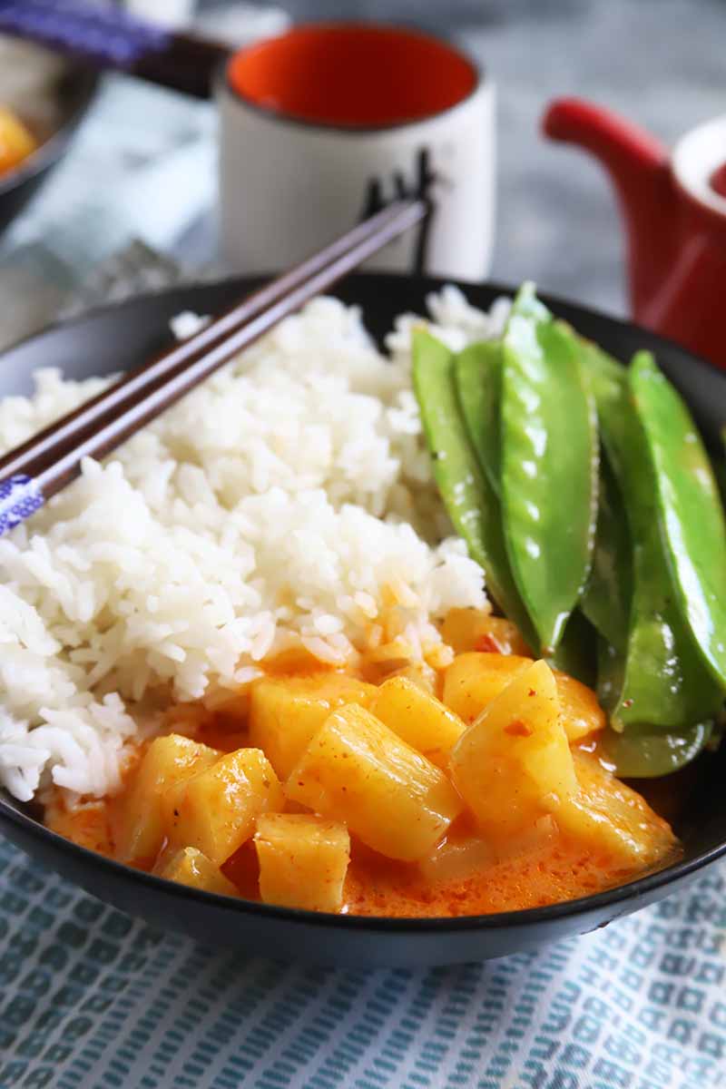 A vertical image of a bowl of Thai pineapple curry served with white rice and snow peas.