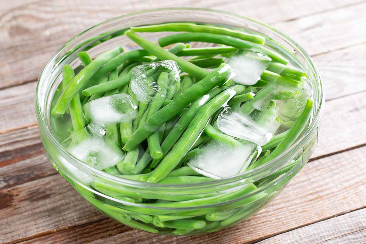 A close up horizontal image of a glass bowl filled with iced water and green beans set on a wooden surface.