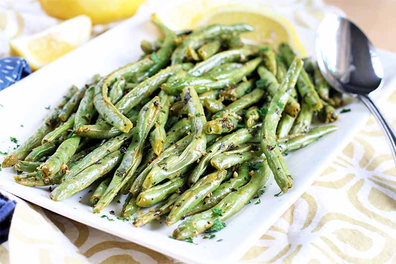 A close up horizontal image of roasted lemon garlic green beans on a white platter with a metal serving spoon to the right of the frame.