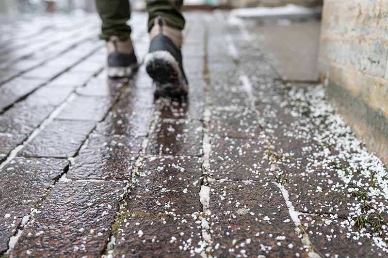 A horizontal image of a person's feet walking down a sidewalk that has had salt applied in winter.
