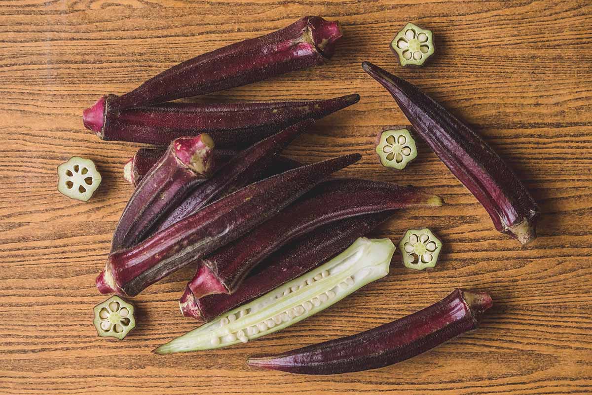A horizontal image of whole and sliced 'Red Burgundy' okra set on a wooden surface.