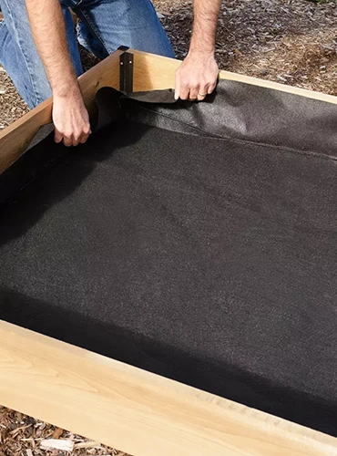 A close up of a gardener applying black liner fabric to a wooden raised bed garden.