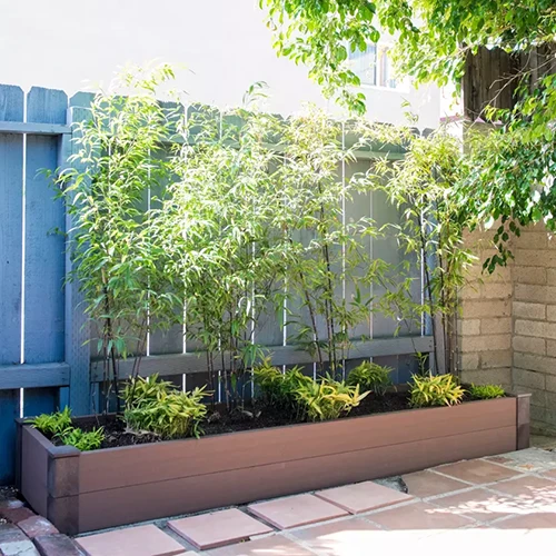 A square image of a recycled composite raised bed set against a blue wooden fence on a patio.