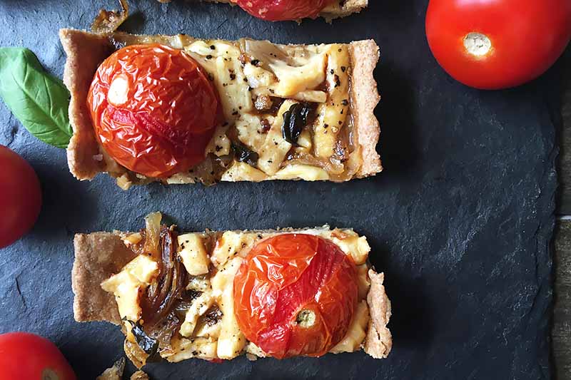 A close up top down image of a tomato tart, fresh out of the oven on a dark slate serving platter.
