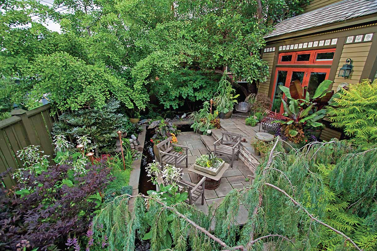 A horizontal image photographed from a slightly elevated position showing a tidy outdoor patio with a variety of potted plants and perennial borders.