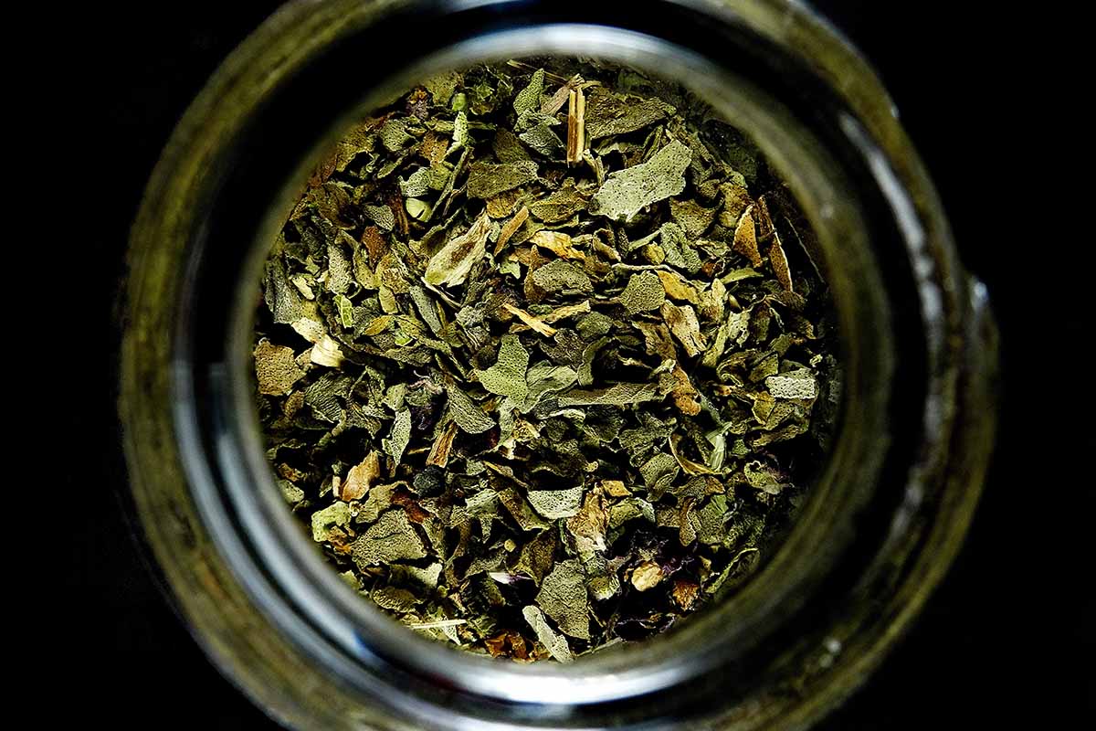 A close up of a jar filled with dried herbs pictured on a dark background.