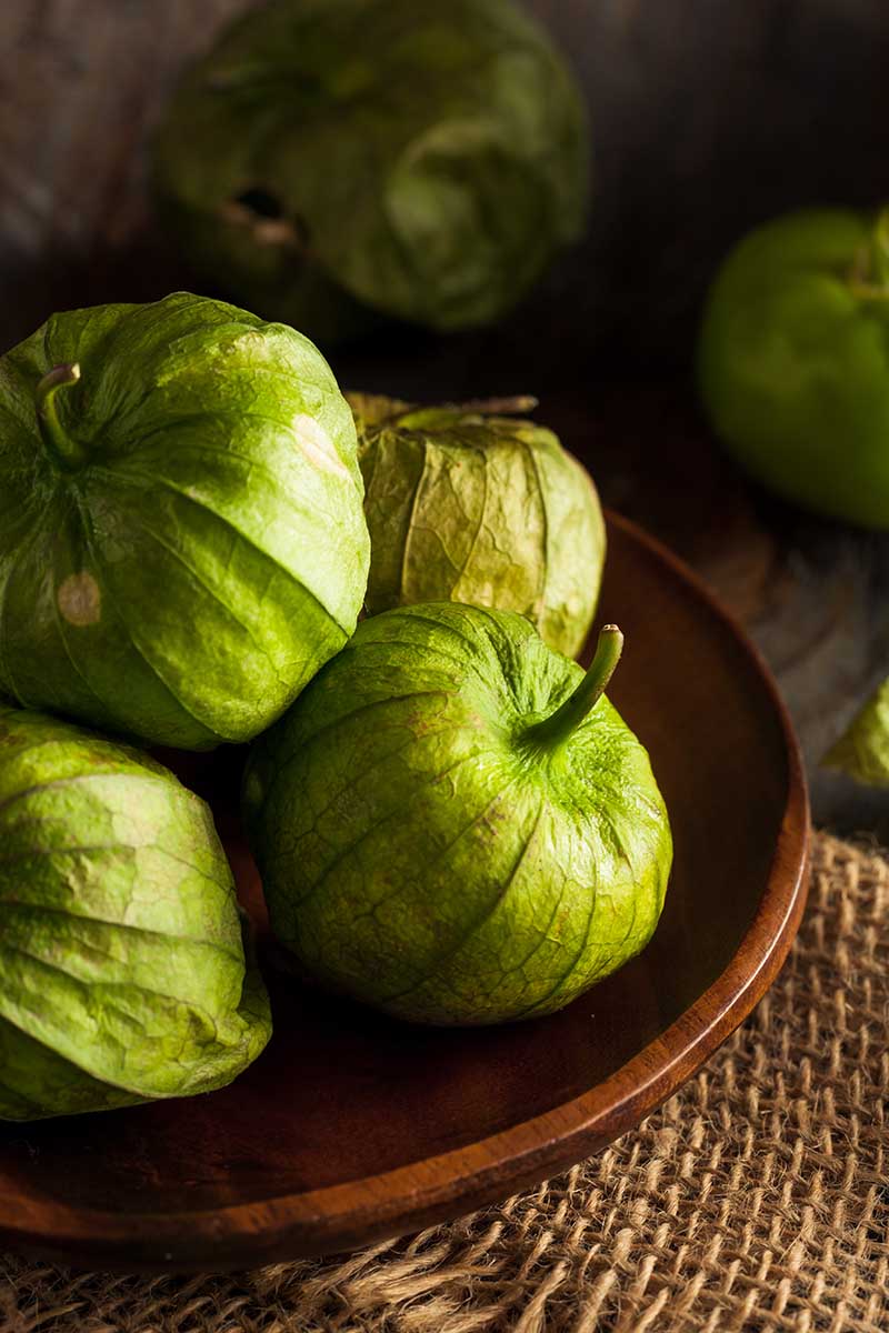 A close up vertical image of freshly harvested tomatillos on a wooden plate pictured on a dark background.
