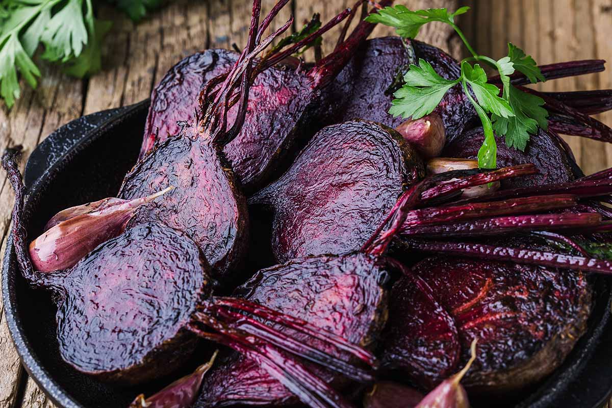 A horizontal image of roasted beets and garlic in a cast iron skillet on a wooden table outdoors.