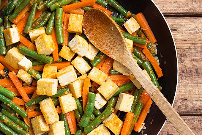 A close up of a tofu and green bean stir fry in a pan set on a wooden surface.