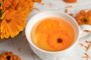 A close up horizontal image of a cup of calendula tea with flowers scattered around on a white wooden surface.