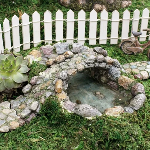 A square image of a Cinda Stone pond and bridge fairy garden with moss, succulents, and a picket fence.