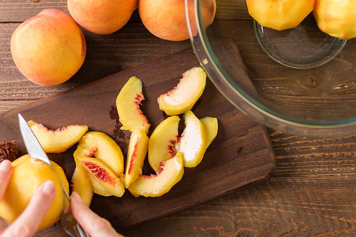 A horizontal image of two hands from the left of the frame slicing peaches on a wooden chopping board.