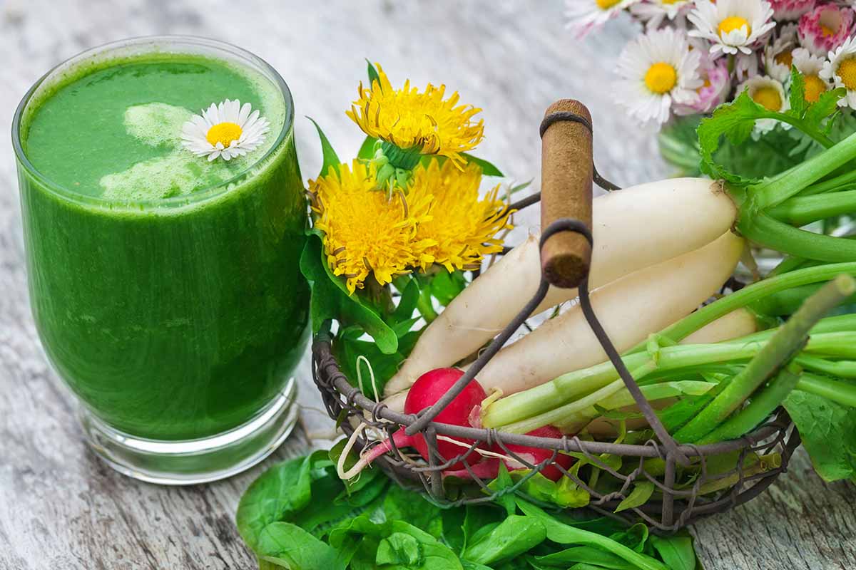 A close up horizontal image of a green smoothie set on a wooden surface with a basket of veggies and flowers next to it.
