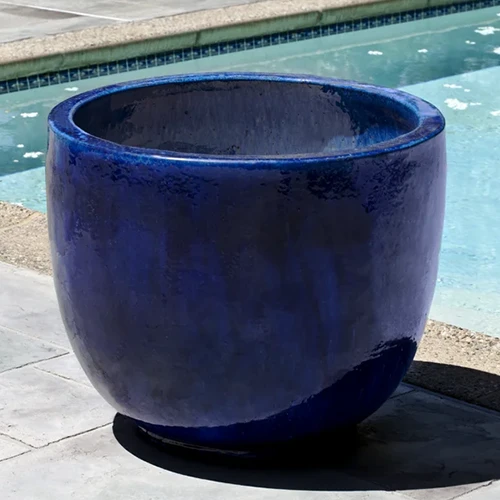 A close up of a blue glazed terra cotta pot set on the ground next to a swimming pool.