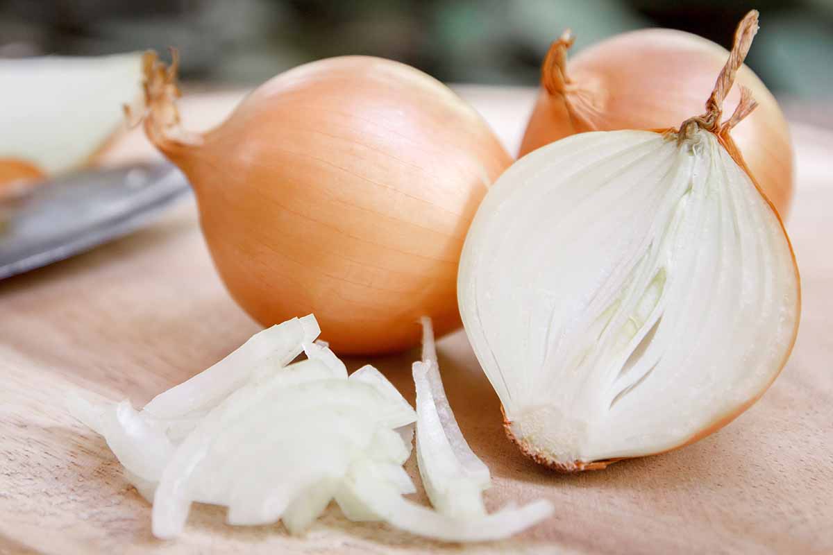 A close up horizontal image of bulb onions whole and sliced on a wooden chopping board.