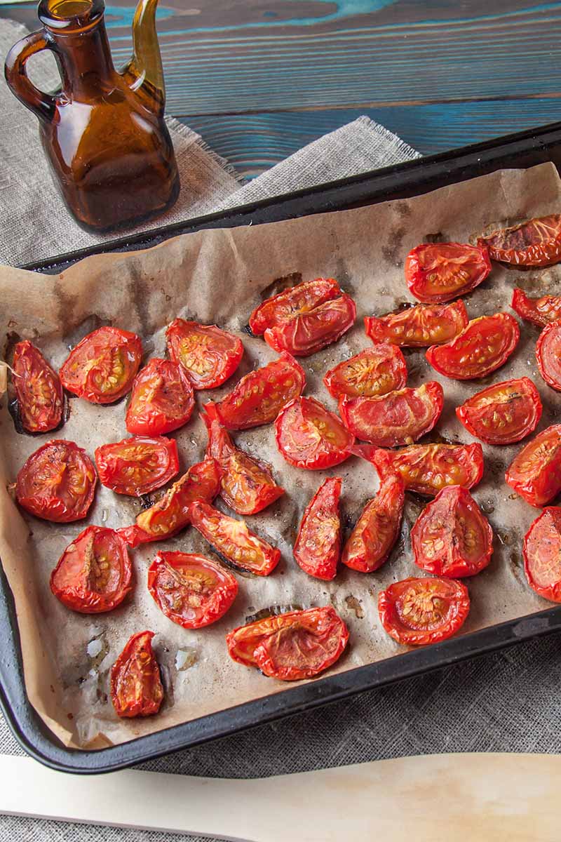 A close up vertical image of a lined baking tray with slices of tomatoes just out of the oven, set on a wooden surface with a vinegar bottle in the background.