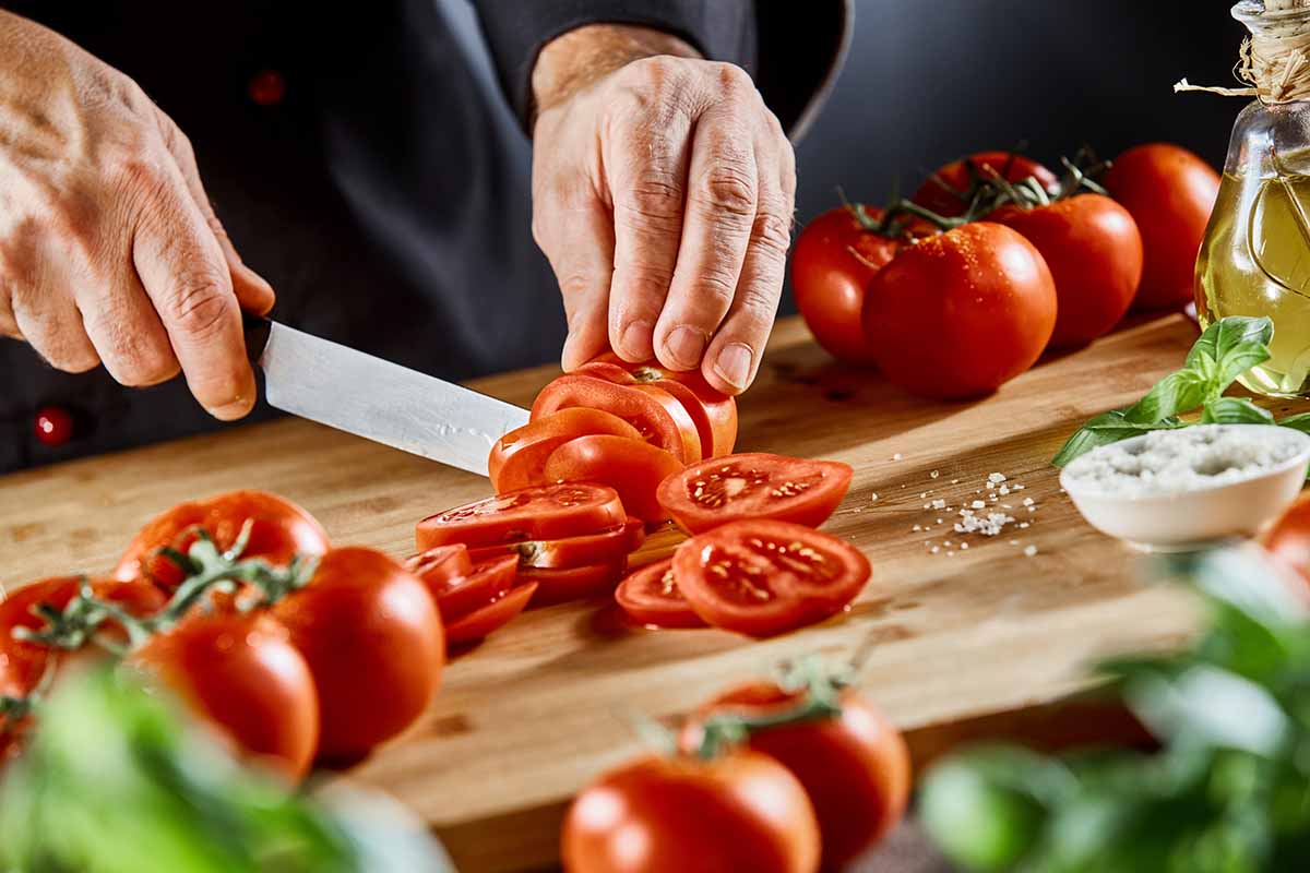 A close up horizontal image of a chef slicing ripe red tomatoes on a wooden chopping board with herbs and salt scattered around.
