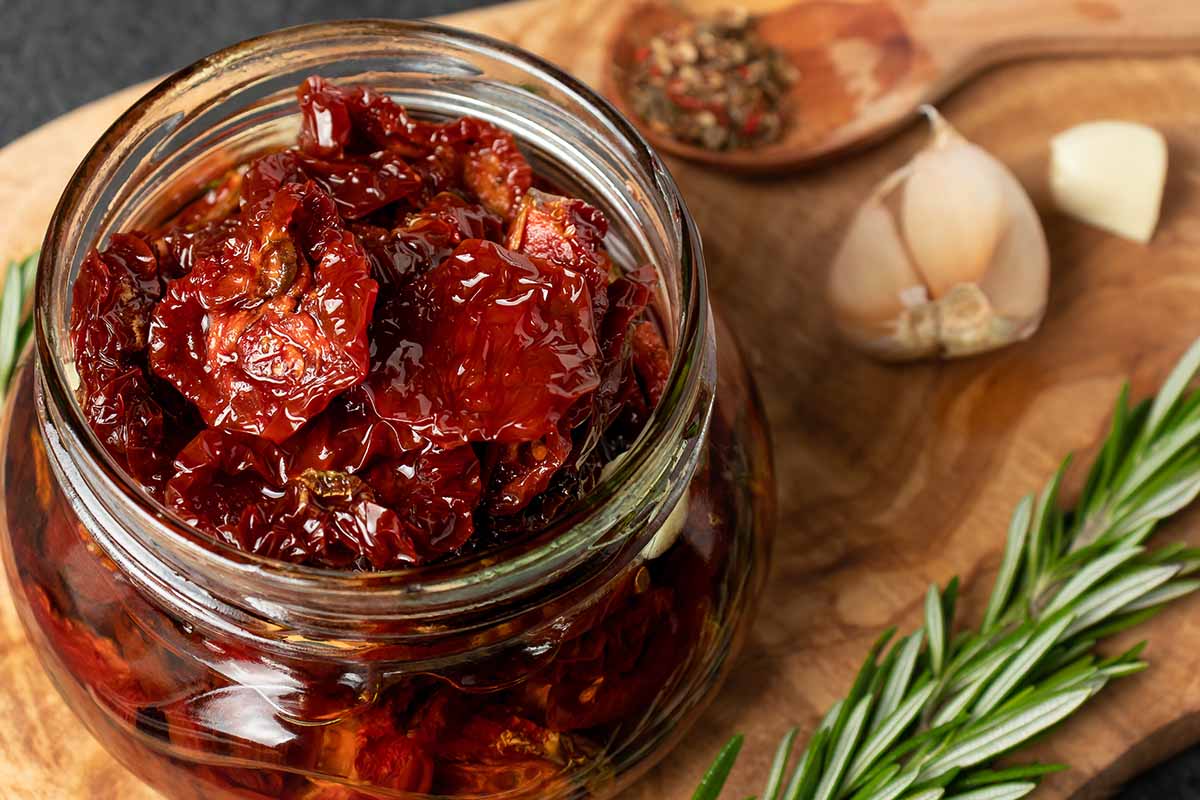 A close up horizontal image of a jar of sun-dried tomatoes in oil set on a wooden surface with garlic and a sprig of rosemary next to it.