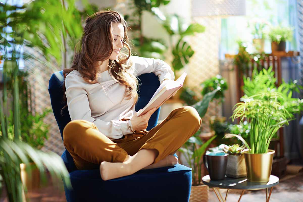 A horizontal image of a woman sitting cross-legged in a chair reading a book, surrounded by houseplants.