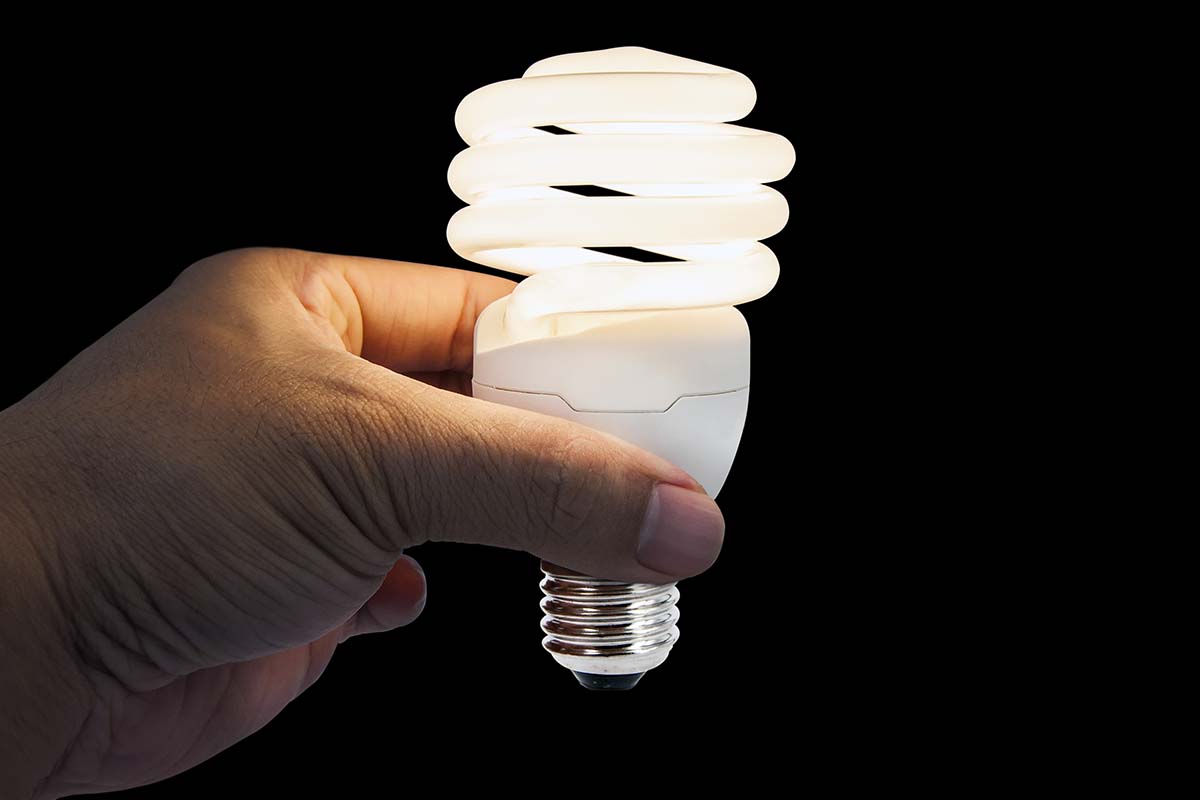 A close up horizontal image of a hand from the left of the frame holding up a light bulb, pictured on a black background.