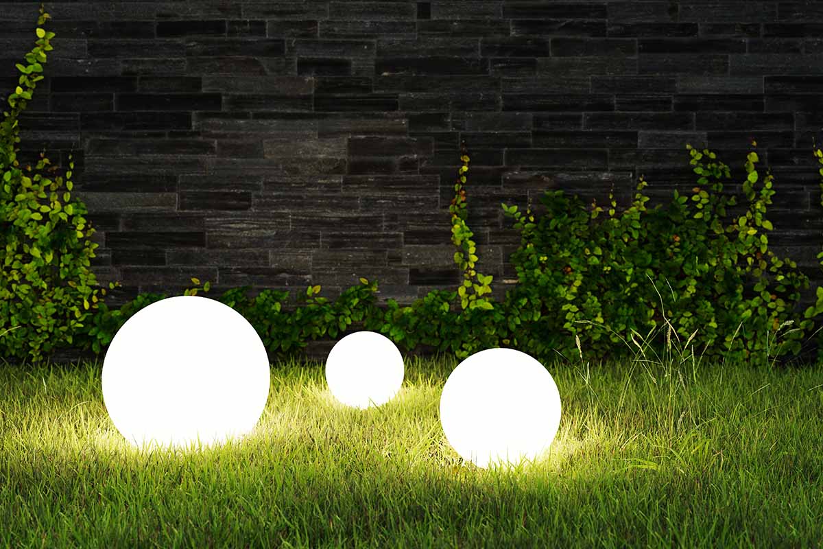 A close up horizontal image of three globe lights set on the lawn with a brick fence in the background.