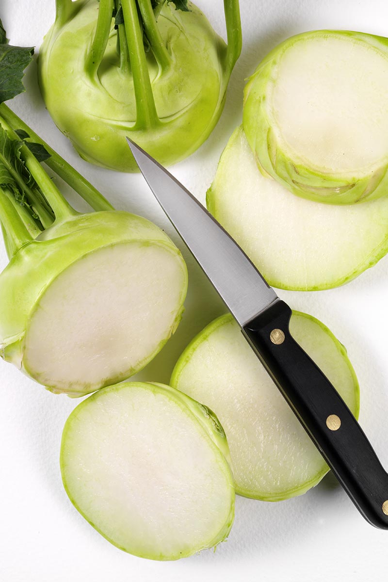 A close up vertical image of whole and sliced kohlrabi set on a white surface with a knife.