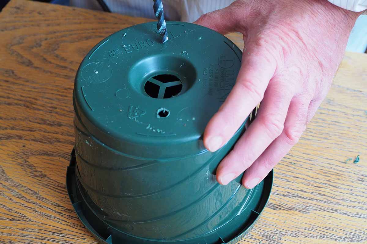 A horizontal close up of a gardener holding a green plastic pot with the bottom facing up. A drill bit is making a hole in the bottom of the pot.