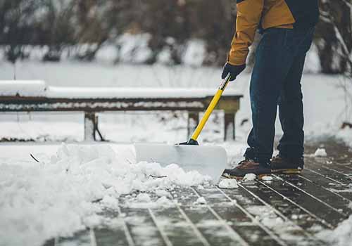 A close up horizontal image of a person shoveling snow on a tiled patio.