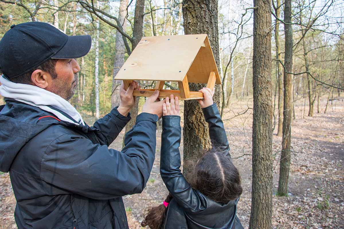 A horizontal photo of a father and daughter installing a wooden bird feeder in a tree.