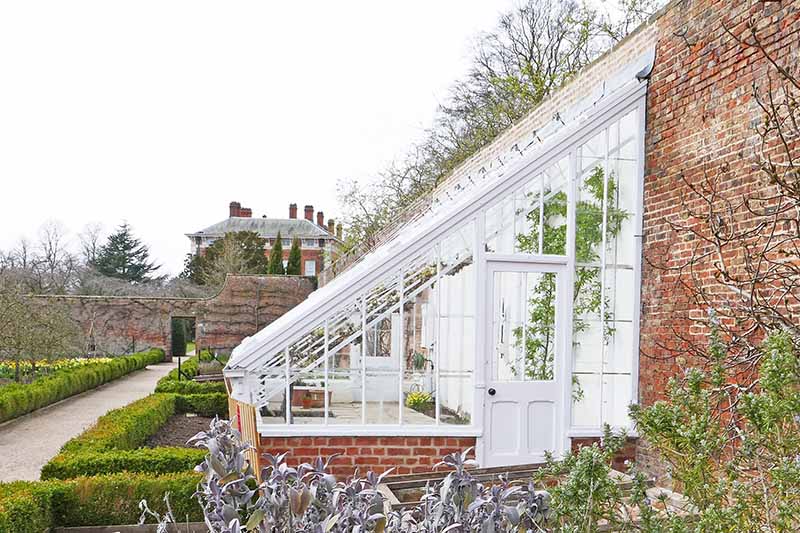 A horizontal image of a lean to glasshouse against a brick wall in a large country garden.