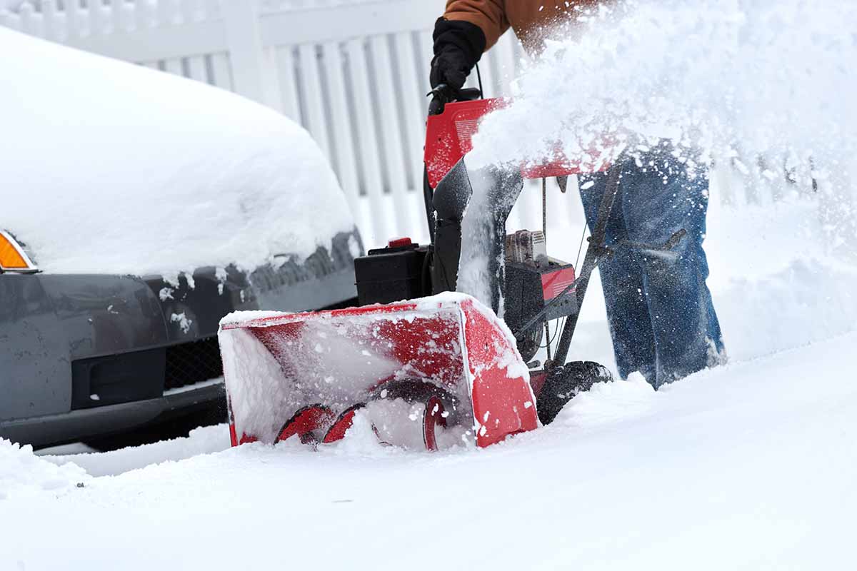 A close up horizontal image of a snowblower clearing a driveway with a car and white fence in soft focus in the background.