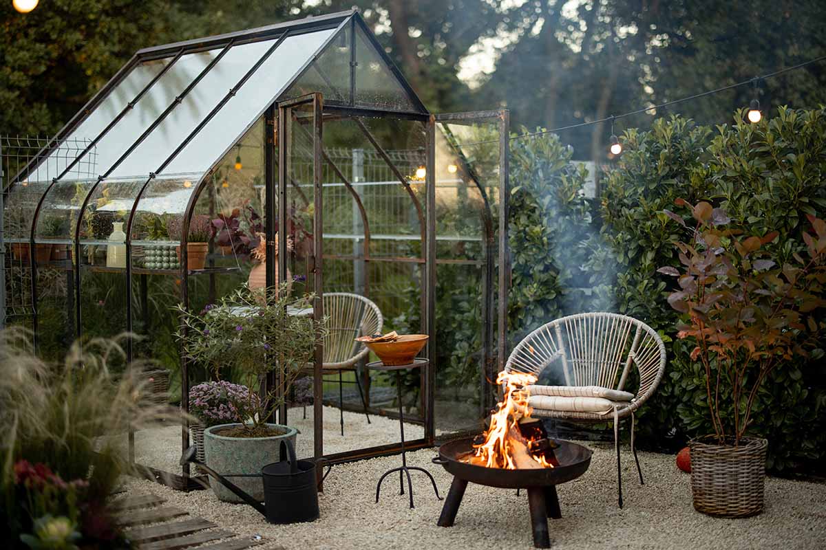 A horizontal image of a greenhouse in the backyard with a seating area and fire pit outside it.