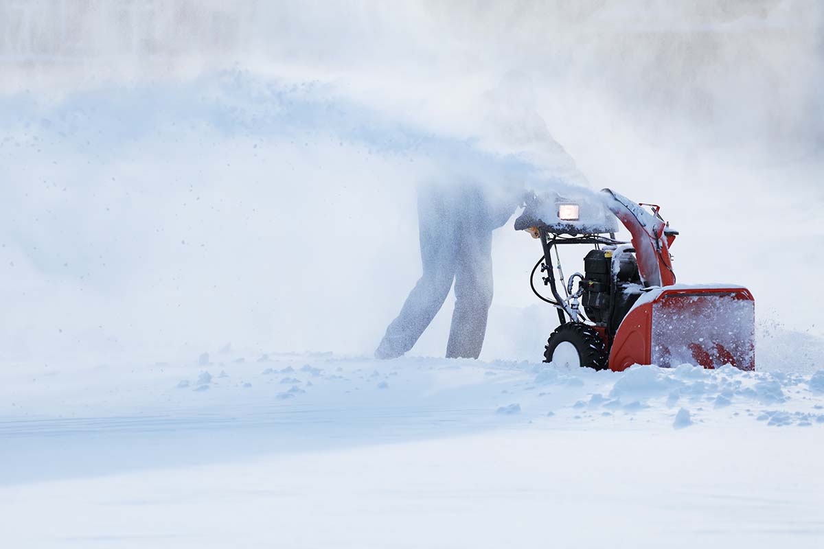 A close up horizontal image of a snowy landscape being cleared with a gas-powered machine pictured on a soft focus background.
