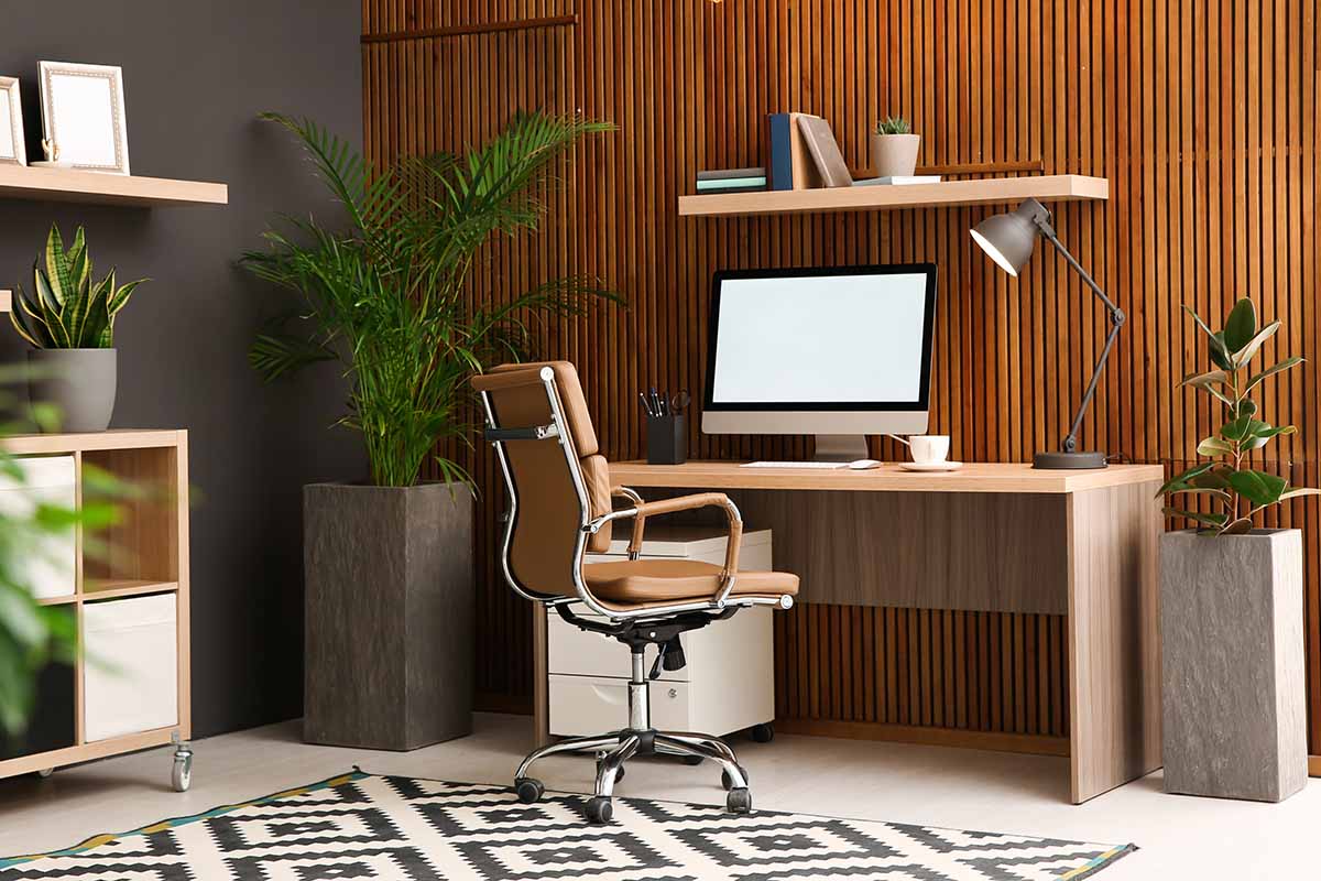A horizontal photo of a home office with a desk against a wooden paneled wall and a large houseplant in the corner.