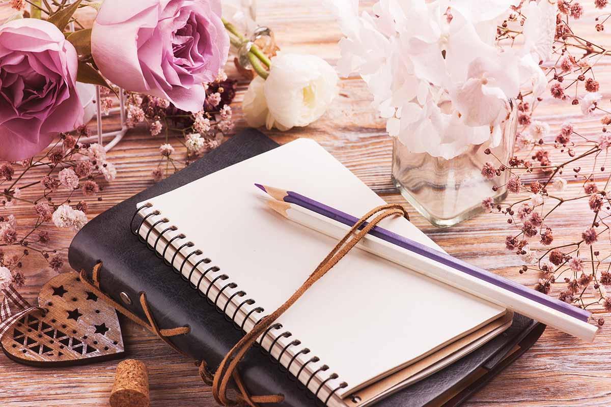 A close up horizontal image of a gardening journal surrounded by flowers set on a wooden surface.