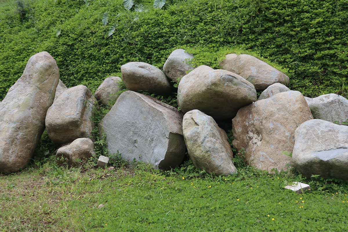 A horizontal image of a pile of large rocks sitting against an outdoor hillside in Indonesia, surrounded by low-growing greenery.