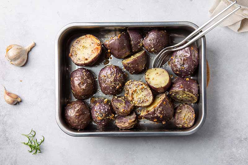 A close up horizontal image of a roasting dish filled with oven baked purple potatoes with herbs and salt set on a marble surface with garlic scattered around.