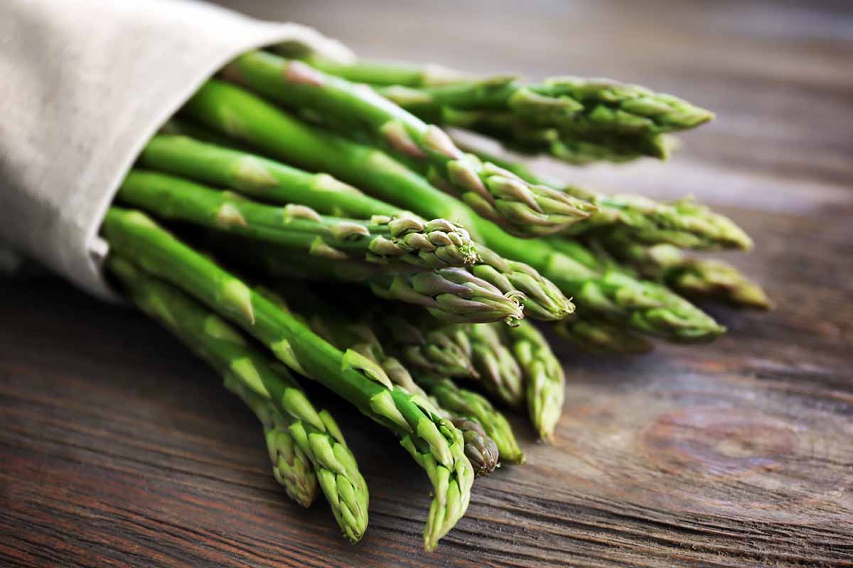 A close up horizontal image of a bunch of asparagus spears set on a wooden table.